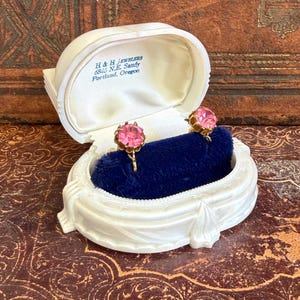 May include: A pair of round, faceted pink stone screw-back earrings set in gold-tone metal, displayed in a white plastic jewelry box with a dark blue velvet insert. The interior lid features the text H & H Jewelers 6845 N.E. Sandy Portland, Oregon.