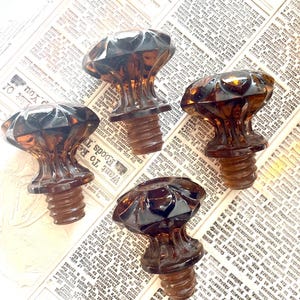 May include: Four amber-colored, faceted glass bottle stoppers with a mushroom-shaped design. The stoppers have a threaded base and are displayed on a vintage newspaper background.