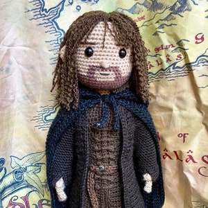 以下が含まれることがあります： 茶色の髪、茶色のひげ、茶色のフード付きマントを着た架空のキャラクターの編みぐるみ人形。人形は茶色のチュニックと茶色のブーツを履いています。人形は地図柄の布地の背景に立っています。