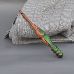 K&ouml;nnte beinhalten: Ein H&auml;kelnadel aus Holz mit einem gr&uuml;nen und braunen Wirbelmuster. Die Nadel liegt auf einer grauen Oberfl&auml;che.