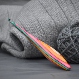 May include: A colorful wooden crochet hook with a pink, green, and orange striped design. The hook is lying on a gray surface next to a ball of gray yarn.