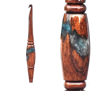 Puede incluir: Un ganchillo artesanal con mango de madera marr&oacute;n oscuro pulido con acabado brillante y una incrustaci&oacute;n central de resina azul y gris. La herramienta presenta una punta c&oacute;nica y una secci&oacute;n de agarre redondeada con detalles torneados.