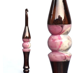 Könnte beinhalten: Ein Häkelnadel aus Holz mit einem pinkfarbenen und weißen Marmormuster. Die Nadel hat einen braunen Griff und eine glatte, polierte Oberfläche.