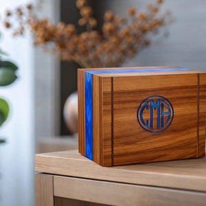 May include: A wooden box with a blue resin inlay and a circular monogram. The box is made of a warm-toned wood and sits on a wooden surface. The monogram features the letters "CMA" in a blue color.