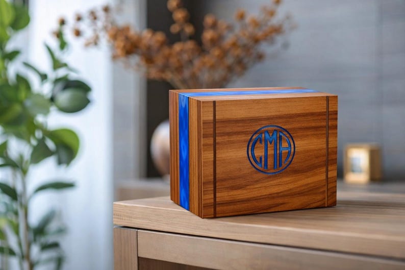 May include: A wooden box with a blue resin inlay and a circular monogram. The box is made of a warm-toned wood and sits on a wooden surface. The monogram features the letters "CMA" in a blue color.