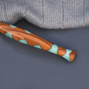 K&ouml;nnte beinhalten: Eine Stricknadel aus Holz mit tealfarbenen und braunen Harzeinlagen. Die Nadel liegt auf einer grauen Oberfl&auml;che.