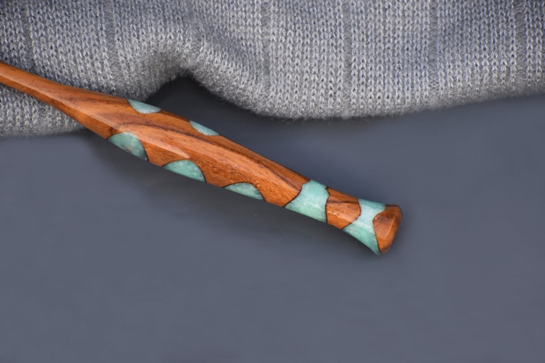 K&ouml;nnte beinhalten: Eine Stricknadel aus Holz mit tealfarbenen und braunen Harzeinlagen. Die Nadel liegt auf einer grauen Oberfl&auml;che.