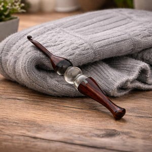 May include: A handcrafted crochet hook with a dark wood handle and a clear, rounded glass center. The hook rests on a folded gray knitted fabric, suggesting a crafting theme. The wooden surface provides a warm contrast.