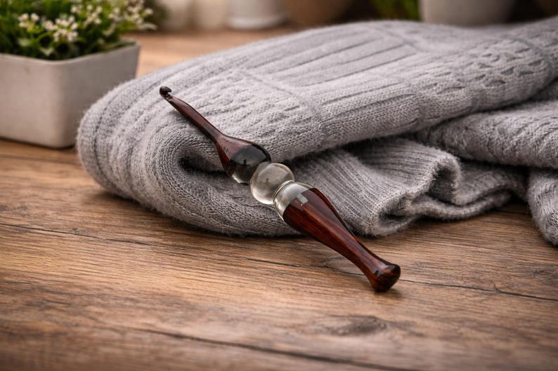 May include: A handcrafted crochet hook with a dark wood handle and a clear, rounded glass center. The hook rests on a folded gray knitted fabric, suggesting a crafting theme. The wooden surface provides a warm contrast.