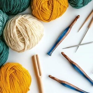 Pu&ograve; includere: Diversi gomitoli di lana nelle tonalit&agrave; verde ottanio, giallo senape e crema sono disposti accanto a tre uncinetti in legno con manici in resina blu e due sottili ferri da maglia in metallo su una superficie bianca.