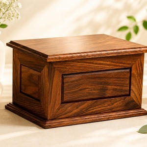 May include: A rectangular wooden cremation urn featuring a rich, dark brown wood grain finish. The urn has a beveled lid, a recessed central panel on the front, and a stepped base design, showcasing a polished surface with visible natural wood patterns.