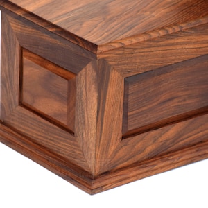 May include: A rectangular wooden box featuring a rich, dark brown grain pattern. The construction includes beveled edges and a recessed panel design on the side, showcasing a smooth, polished finish with visible natural wood textures throughout.