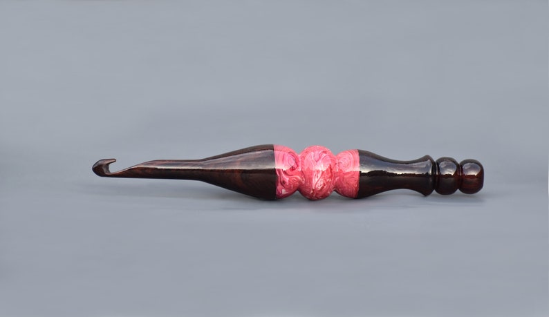 May include: A wooden crochet hook with a dark brown handle and a pink and white marbled section in the middle.