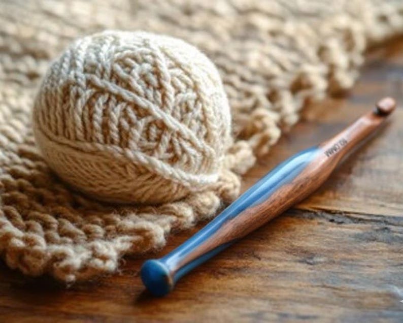 Pu&ograve; includere: Un gomitolo di lana color crema con un motivo a maglia testurizzato e un uncinetto con manico in legno lucido e inserto in resina blu, chiaramente contrassegnato con la misura 10.0MM, poggiato vicino a un pezzo di tessuto a maglia abbinato.