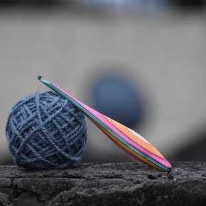 May include: A ball of blue yarn and a colorful wooden crochet hook on a gray textured surface.