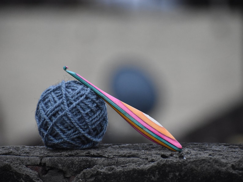 May include: A ball of blue yarn and a colorful wooden crochet hook on a gray textured surface.
