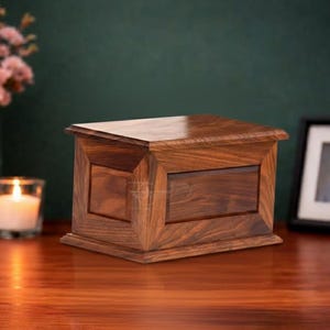 May include: A rectangular wooden cremation urn featuring a rich brown grain pattern, a flat top lid, and a beveled frame design on the front panel. The box is constructed with smooth, polished wood surfaces and a sturdy base, showcasing natural wood textures.