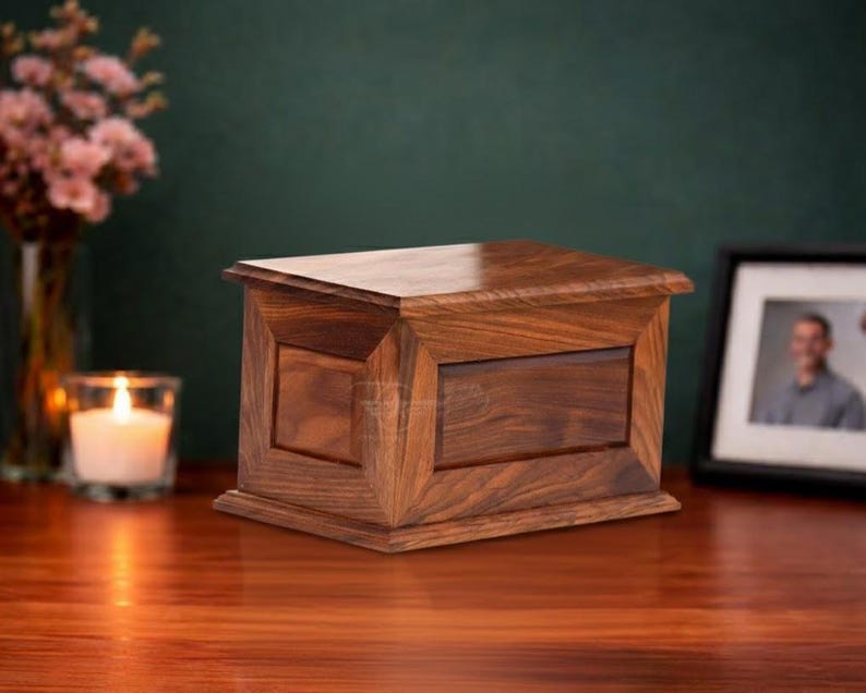 May include: A rectangular wooden cremation urn featuring a rich brown grain pattern, a flat top lid, and a beveled frame design on the front panel. The box is constructed with smooth, polished wood surfaces and a sturdy base, showcasing natural wood textures.