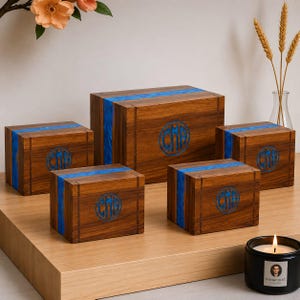 May include: Five wooden boxes of varying sizes with a dark wood grain finish. Each box features a vibrant blue resin stripe and a circular monogram. The boxes are arranged on a light wood surface, with a lit candle in the foreground.