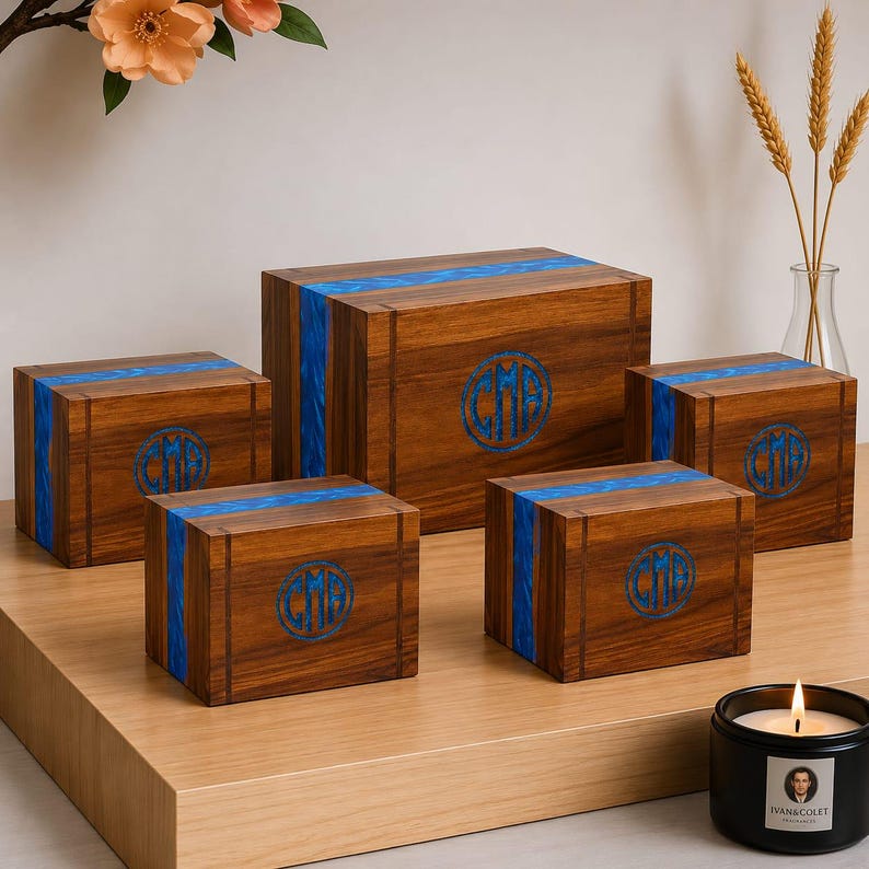May include: Five wooden boxes of varying sizes with a dark wood grain finish. Each box features a vibrant blue resin stripe and a circular monogram. The boxes are arranged on a light wood surface, with a lit candle in the foreground.