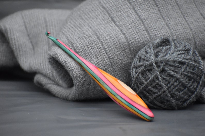 May include: A colorful wooden crochet hook with a pink, green, and orange striped design. The hook is lying on a gray surface next to a ball of gray yarn.