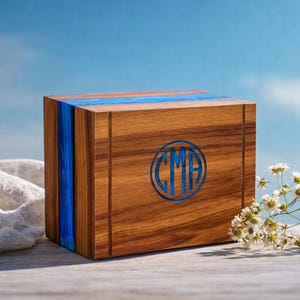 May include: A wooden box with a blue stripe and blue initials. The box is made of wood with a natural grain and a blue resin stripe. The initials "CMA" are in a blue circle on the front. The background is a blue sky.