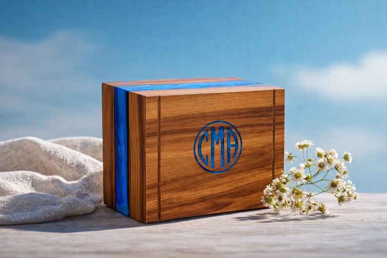 May include: A wooden box with a blue stripe and blue initials. The box is made of wood with a natural grain and a blue resin stripe. The initials "CMA" are in a blue circle on the front. The background is a blue sky.