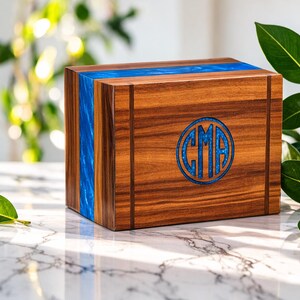 May include: A wooden box with a blue resin inlay and a circular monogram. The box is made of dark wood with vertical grain and a blue resin stripe. The monogram features the letters "CMA" in a blue design.