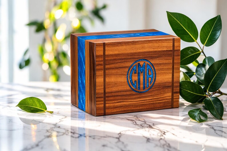 May include: A wooden box with a blue resin inlay and a circular monogram. The box is made of dark wood with vertical grain and a blue resin stripe. The monogram features the letters "CMA" in a blue design.