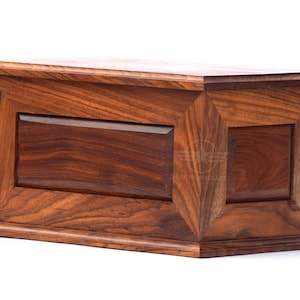 May include: A rectangular wooden box with a rich brown grain pattern, featuring a beveled lid and a base with stepped edges. The front panel has a recessed center section framed by angled wood trim, showcasing a natural polished finish throughout.