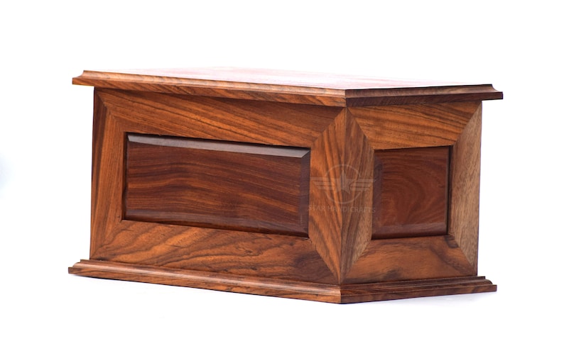 May include: A rectangular wooden box with a rich brown grain pattern, featuring a beveled lid and a base with stepped edges. The front panel has a recessed center section framed by angled wood trim, showcasing a natural polished finish throughout.