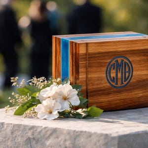 May include: A wooden box with a blue stripe and a circular monogram. The box is made of wood and has a natural finish. White flowers and greenery are placed in front of the box.