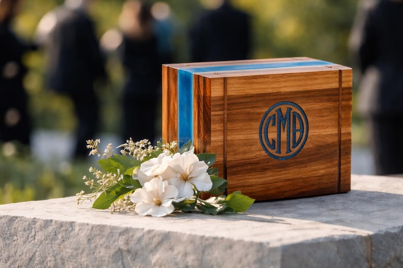 May include: A wooden box with a blue stripe and a circular monogram. The box is made of wood and has a natural finish. White flowers and greenery are placed in front of the box.