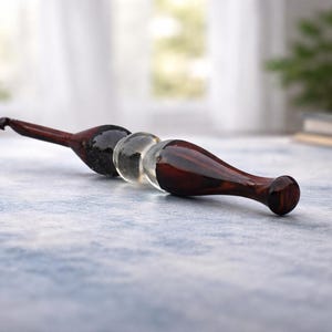 May include: A wooden massage tool with a dark brown finish and a clear glass sphere in the center. The tool is designed with a curved handle and a rounded end. The background is blurred, suggesting a spa or wellness setting.