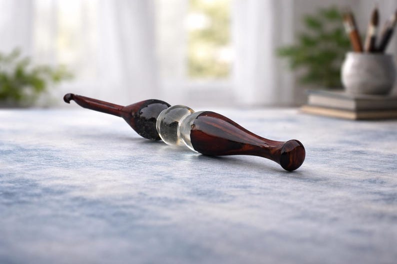 May include: A wooden massage tool with a dark brown finish and a clear glass sphere in the center. The tool is designed with a curved handle and a rounded end. The background is blurred, suggesting a spa or wellness setting.