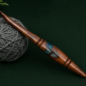 Puede incluir: Un ganchillo artesanal con mango de madera pulida en color marr&oacute;n oscuro y una incrustaci&oacute;n central de resina azul transl&uacute;cida con remolinos. El ganchillo est&aacute; colocado en diagonal junto a un ovillo de lana gris sobre una superficie verde.