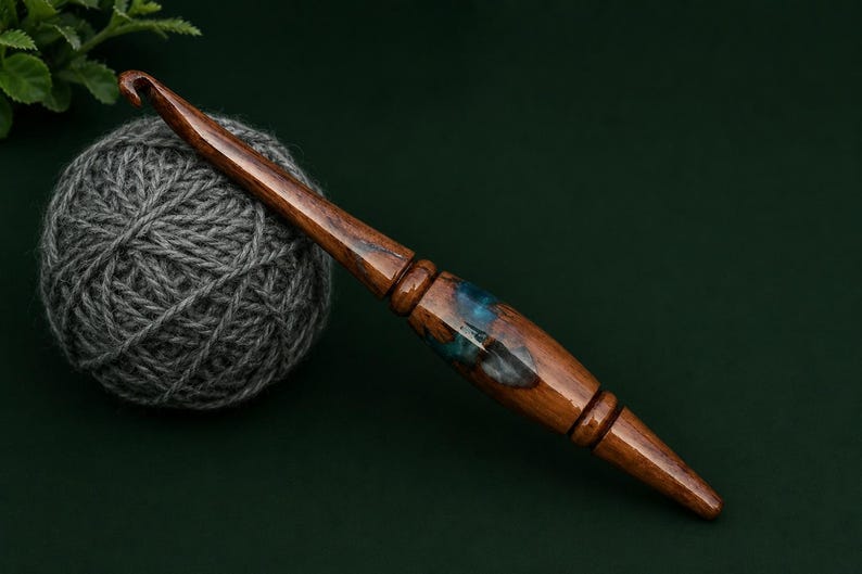 Puede incluir: Un ganchillo artesanal con mango de madera pulida en color marr&oacute;n oscuro y una incrustaci&oacute;n central de resina azul transl&uacute;cida con remolinos. El ganchillo est&aacute; colocado en diagonal junto a un ovillo de lana gris sobre una superficie verde.