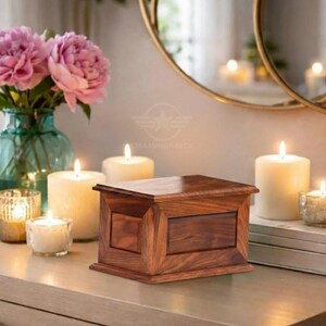 May include: A rectangular wooden box with a polished finish and visible grain patterns, featuring a flat lid and a central panel design on the front. The box is positioned among several lit pillar and votive candles and a glass vase with pink flowers.