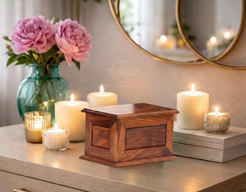 May include: A rectangular wooden box with a polished finish and visible grain patterns, featuring a flat lid and a central panel design on the front. The box is positioned among several lit pillar and votive candles and a glass vase with pink flowers.