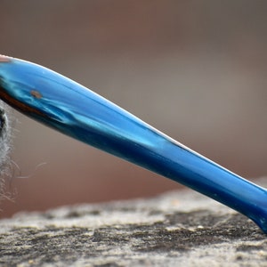 Pu&ograve; includere: Un uncinetto con un manico realizzato in una combinazione di legno marrone scuro e resina blu perlata, liscia e lucida. L'uncinetto si assottiglia verso l'estremit&agrave;, mostrando una netta transizione tra la venatura del legno e il colore blu.