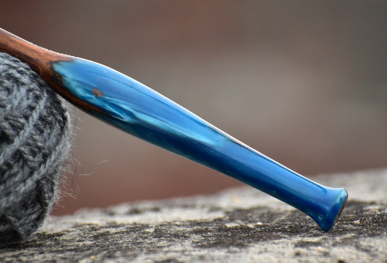 Pu&ograve; includere: Un uncinetto con un manico realizzato in una combinazione di legno marrone scuro e resina blu perlata, liscia e lucida. L'uncinetto si assottiglia verso l'estremit&agrave;, mostrando una netta transizione tra la venatura del legno e il colore blu.