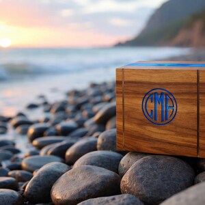 May include: A wooden box with a blue inlay and a circular monogram. The box is made of a dark wood and sits on a rocky beach with the ocean and a sunset in the background. The monogram features the letters "CMF".