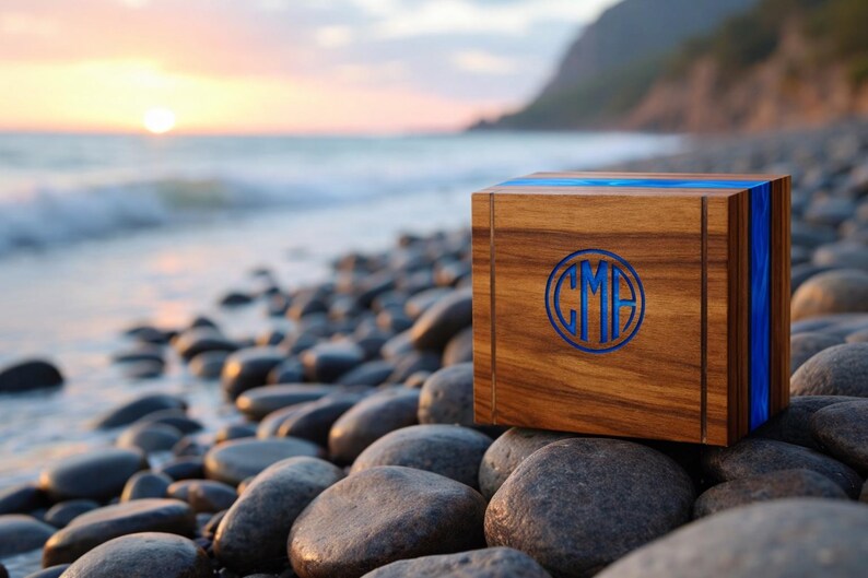 May include: A wooden box with a blue inlay and a circular monogram. The box is made of a dark wood and sits on a rocky beach with the ocean and a sunset in the background. The monogram features the letters "CMF".
