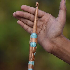 K&ouml;nnte beinhalten: Ein H&auml;kelnadel aus Holz mit einem blauen und wei&szlig;en Design. Die Nadel wird in der Hand einer Person gehalten.