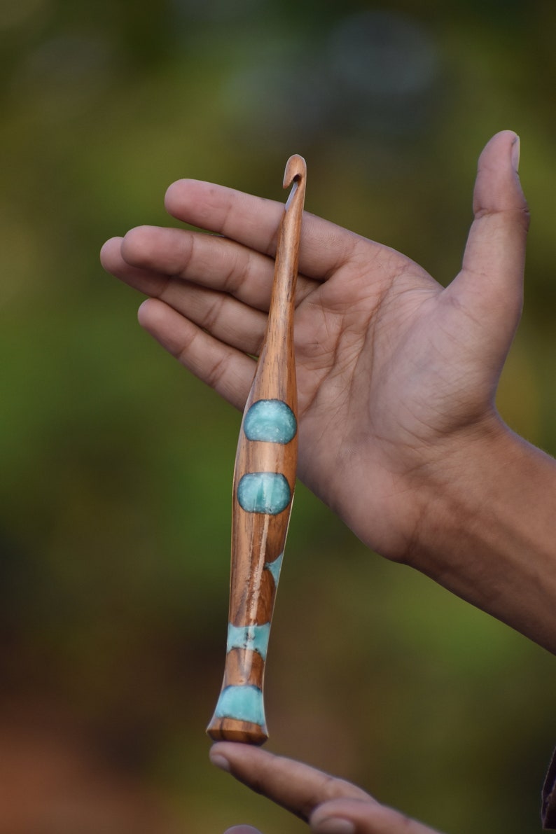 K&ouml;nnte beinhalten: Ein H&auml;kelnadel aus Holz mit einem blauen und wei&szlig;en Design. Die Nadel wird in der Hand einer Person gehalten.