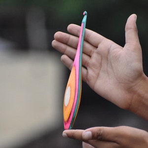 May include: A colorful crochet hook with a pink, orange, and teal design. The hook is made of wood and has a smooth finish.