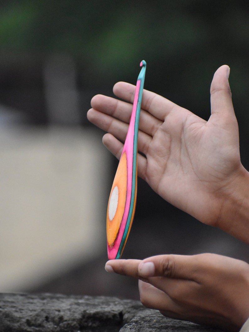 May include: A colorful crochet hook with a pink, orange, and teal design. The hook is made of wood and has a smooth finish.