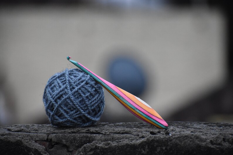 May include: A ball of dark blue yarn and a colorful wooden crochet hook on a gray textured surface.