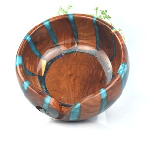 Pu&ograve; includere: Una ciotola per lana rotonda in legno con finitura lucida color marrone e strisce verticali intarsiate in resina turchese traslucida. La ciotola presenta un interno liscio e curvo e un'apertura a spirale sul lato per il filo.