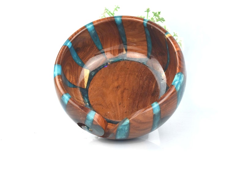 Pu&ograve; includere: Una ciotola per lana rotonda in legno con finitura lucida color marrone e strisce verticali intarsiate in resina turchese traslucida. La ciotola presenta un interno liscio e curvo e un'apertura a spirale sul lato per il filo.